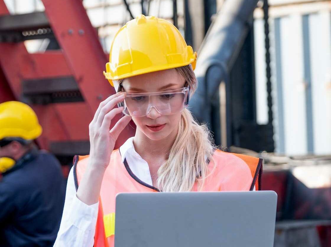 Pretty foreman woman or worker stand with touch her safety eye glasses and holding laptop